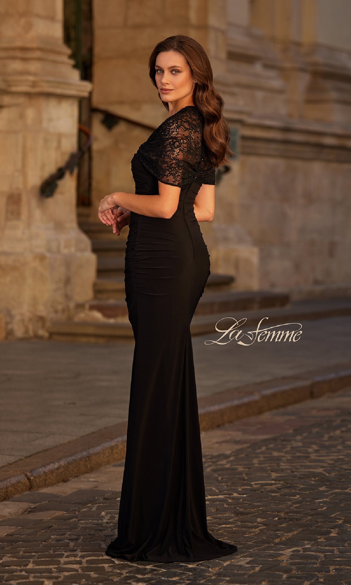 A woman wearing the Lace-Sleeve Black Formal Dress: La Femme 33133 stands on a cobblestone street near a stone building, looking over her shoulder.